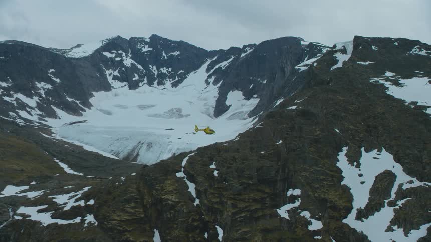 A helicopter flies over a snowy mountain range and towards a river