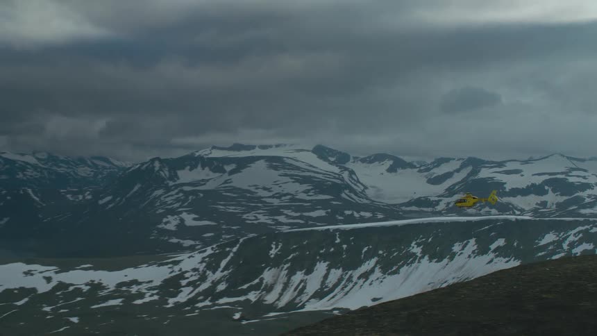 A yellow and red rescue helicopter is flying over a mountain ridge with snow covered mountains in the distance