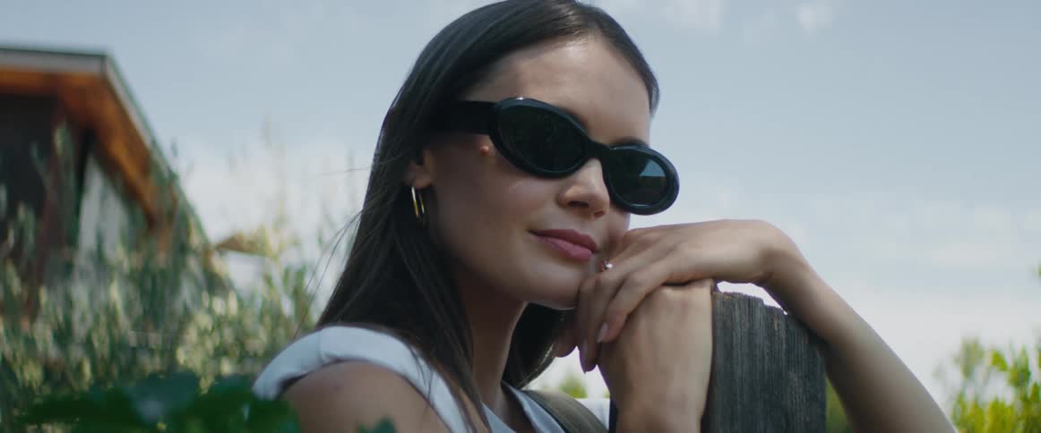 A young woman with long dark hair and wearing sunglasses is outside leaning on a post and looking around