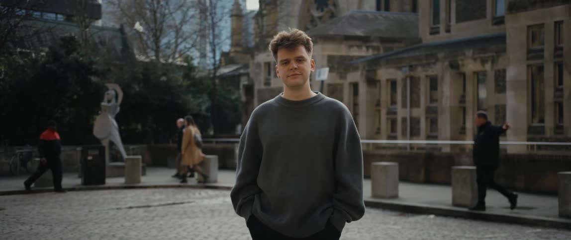 Man poses in courtyard as pedestrians cross past statue and buildings in background