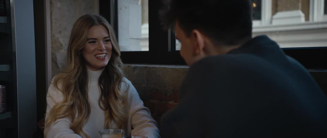 Blonde woman with white sweater and wavy hair sits at table with boyfriend at cafe smiling and talking to each other