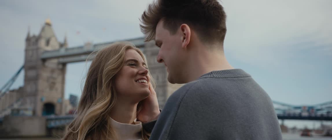 A man in front of a bridge leans forwards and kisses a woman on the forehead