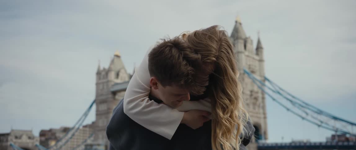 Woman hugs man giving her a piggyback ride and kisses his cheek with landmark bridge in the background