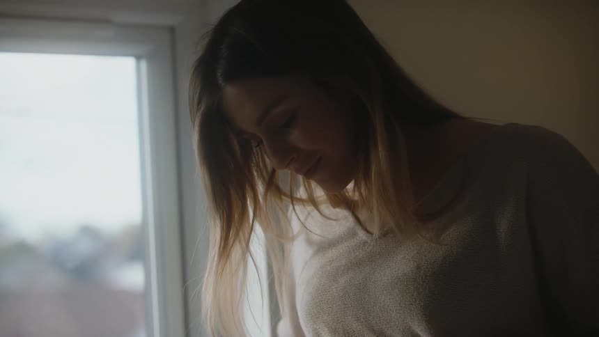 A pregnant woman caresses her belly while standing near a window indoors during the day