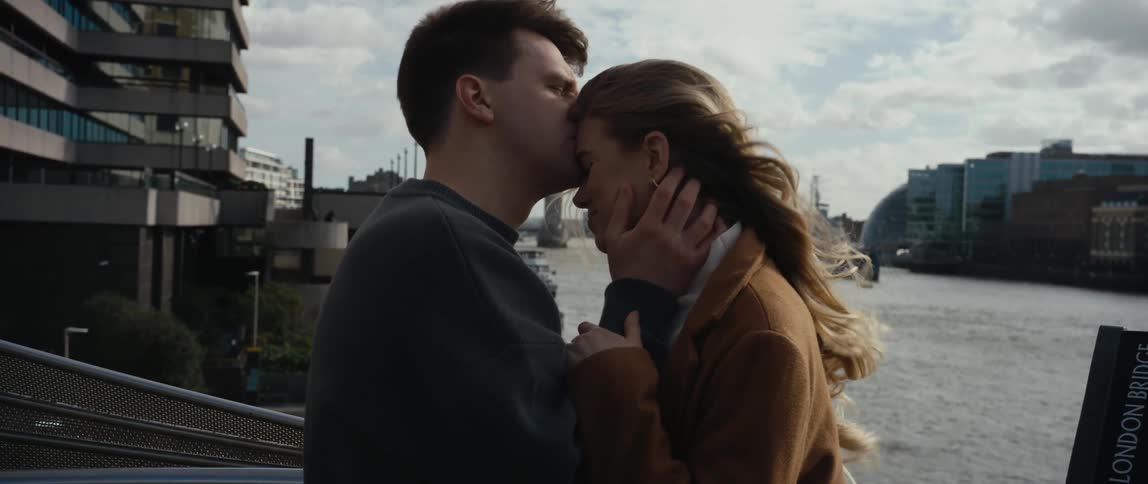 Man kisses woman on forehead as they stand near river with landmark bridge in the distance