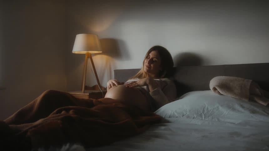 Pregnant woman with brown hair lies on bed rubs her belly in bedroom