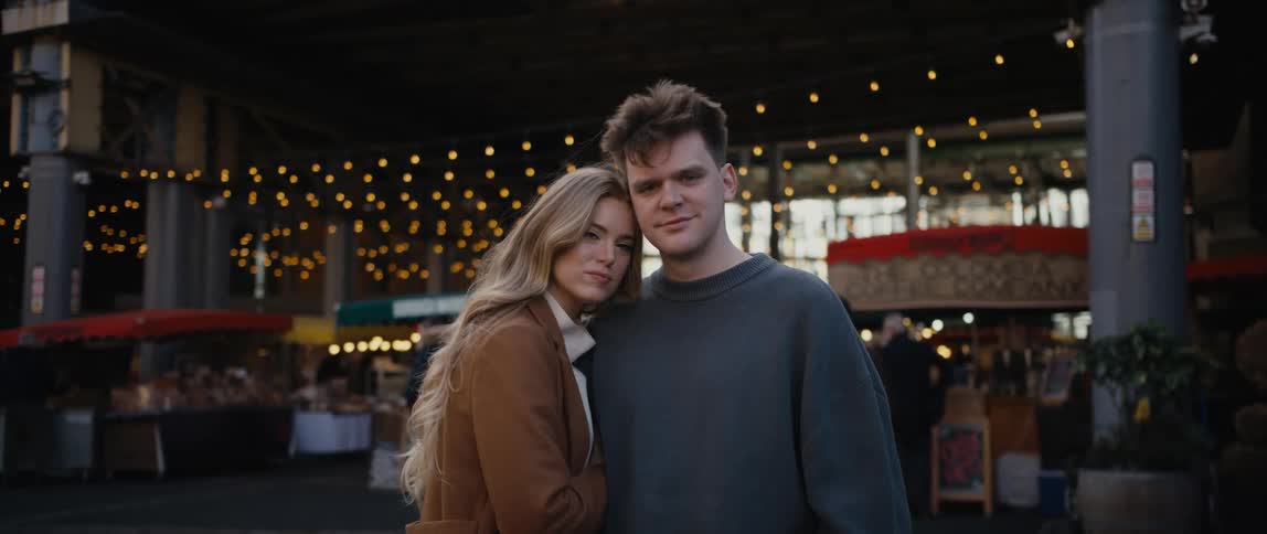A man and woman look at one another before turning to look into the camera while standing outdoors during the day