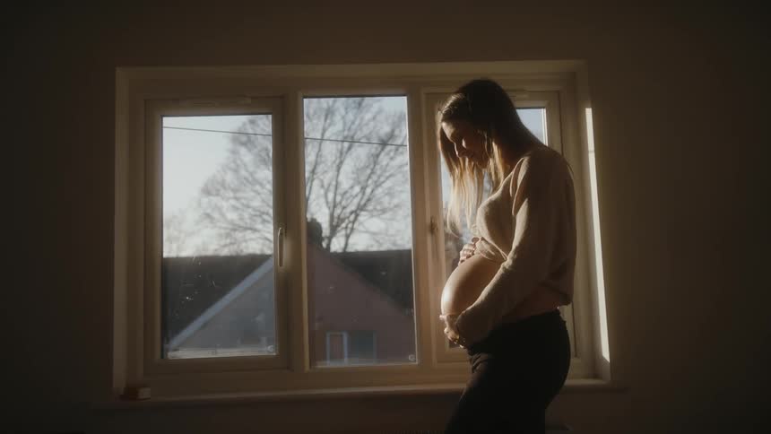 Pregnant woman standing in front of a window inside the house and gently running her hand through her belly