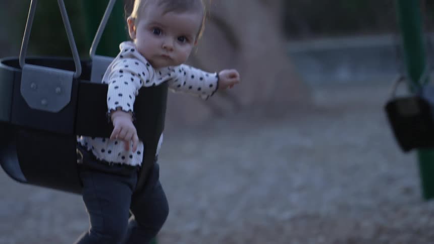 Mother swings baby in swing seat in playground at sunset
