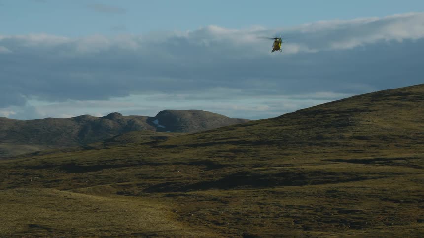 A yellow and red rescue helicopter is flying low over hills with mountains in the distance