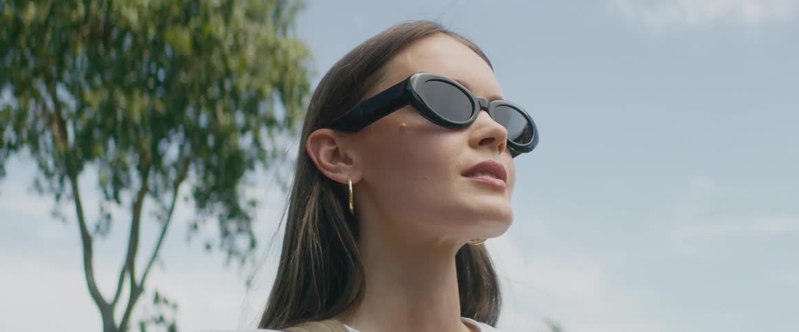 A young woman with long dark hair and wearing sunglasses is outside and the wind is blowing her hair