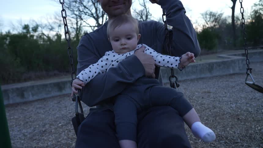 Father sitting at the swing holding his baby in his lap and slowly swinging back and forth