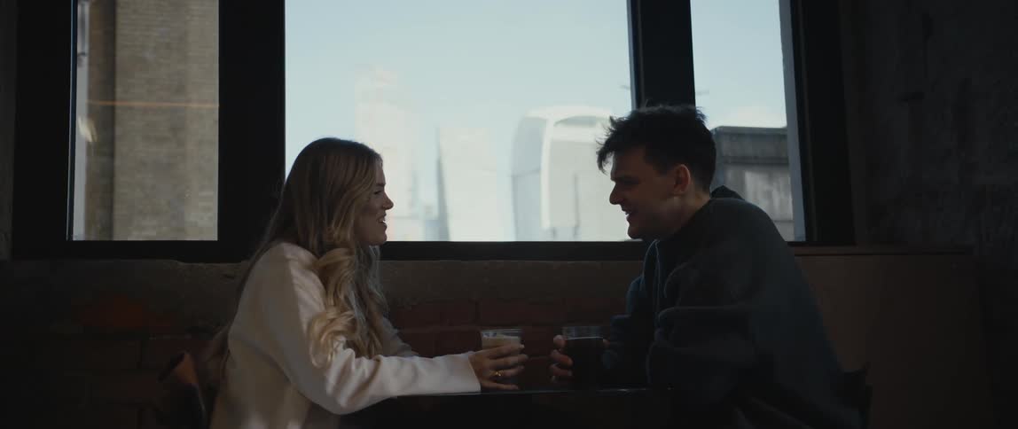 Couple with drinks sit at a small table near a window showing buildings