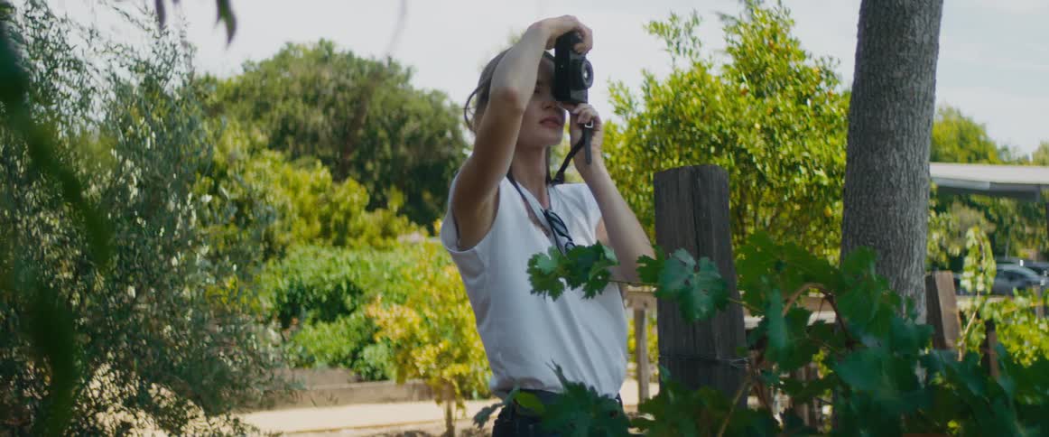 A young woman with long dark hair is outside taking pictures with a camera