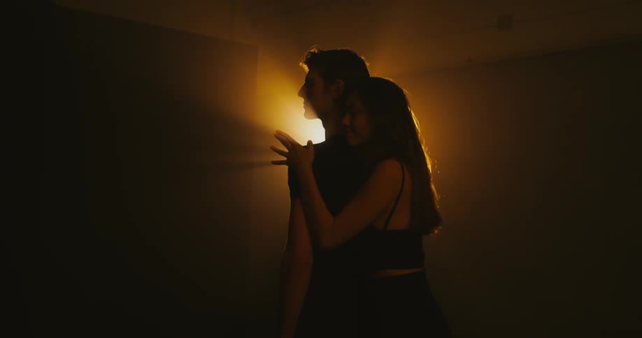 Man standing alone in an empty dark room while a woman approaches and hugs him from behind