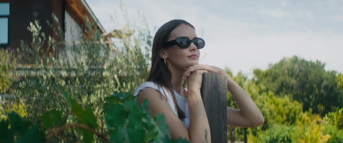 A young woman with long dark hair and wearing sunglasses is outside leaning on a post and looking around