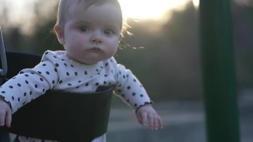 Baby swinging on a fullbucket swing seat at an outdoors playground with an adult person taking care of it