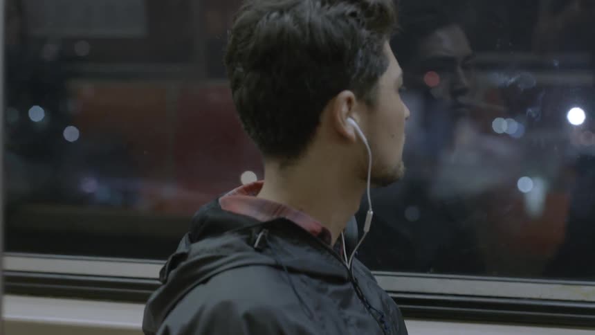 Man sitting on a bus at night looking outside the window with earbuds in