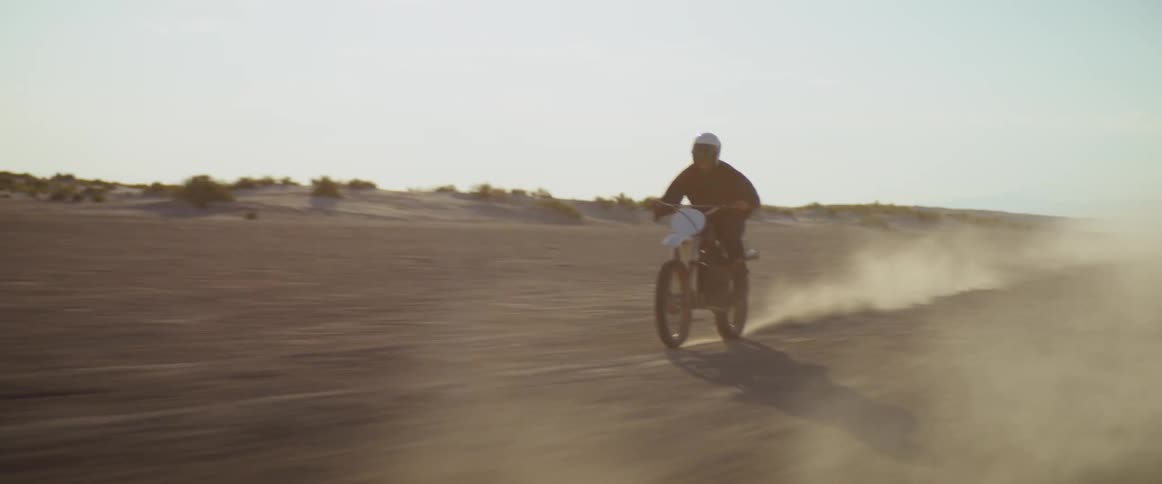 A man wearing a white helmet drives his motorcycle across a dusty desert plain
