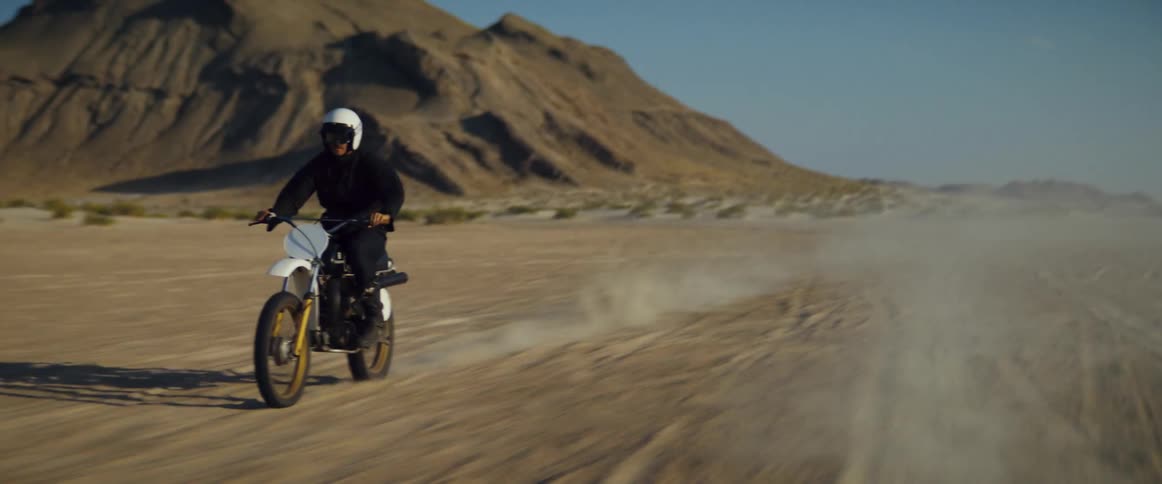 A cyclist wearing a helmet is riding a motorcycle across the desert with mountains in the distance
