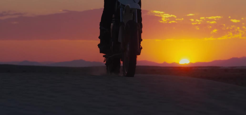 Motorcyclist rides over sand dune as sun sitting on horizon over mountains colors the sky orange and red