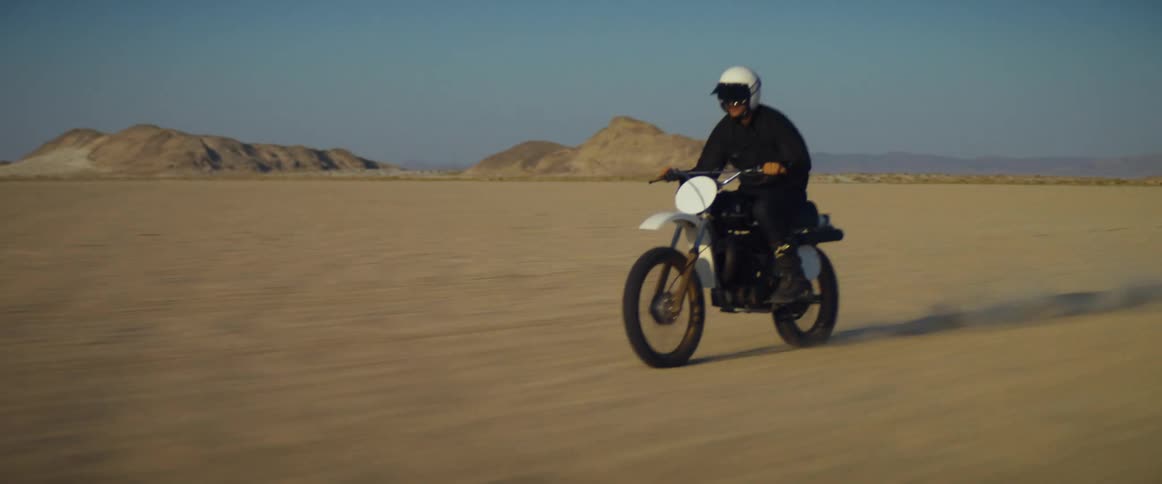 A cyclist wearing a helmet is riding a motorcycle across the desert with mountains in the distance