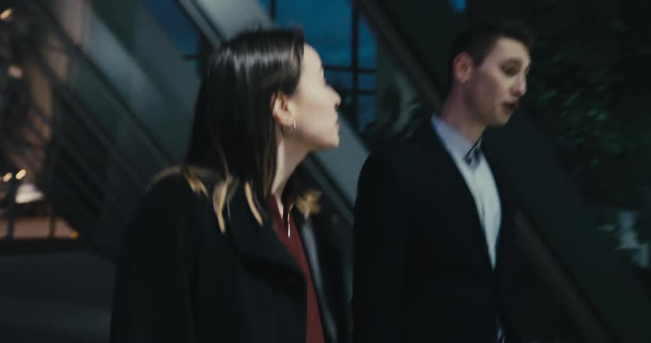 A woman and man walk through a city together while talking at night before the woman grabs the arm of the man to stop him and walks off