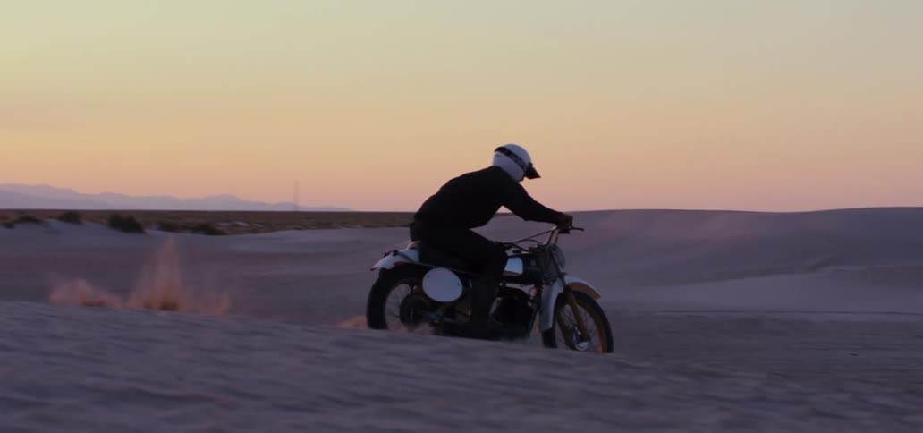 A man wearing a white helmet drives his motorcycle through a desert and completes a jump