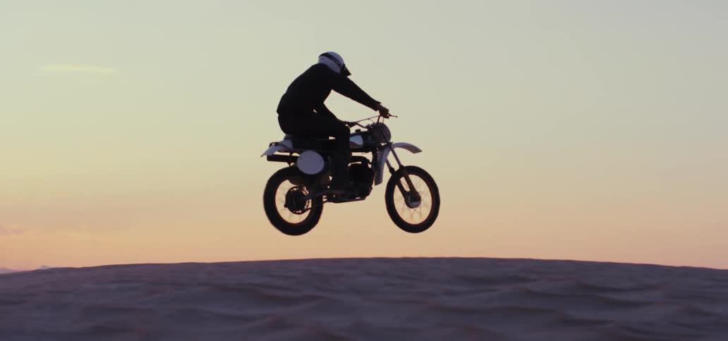 Male biker with white helmet black jacket and pants rides dirt bike and jumps off sand dune at sunset