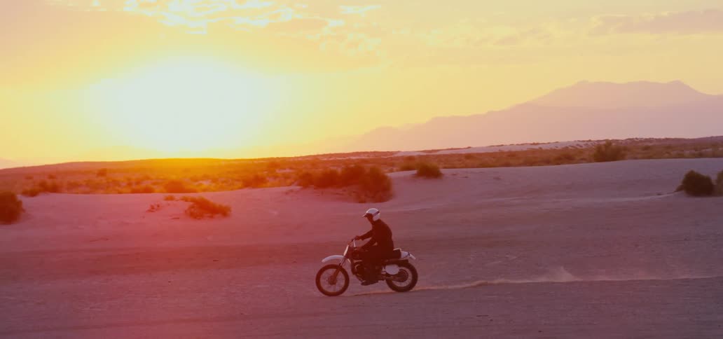 Man with white helmet black jacket and pants rides dirt bike in the desert at sunset