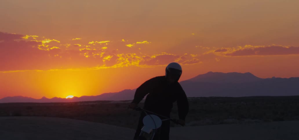 A cyclist wearing a helmet is riding a motorcycle in the desert with silhouettes of mountains in the distance and the sun on the horizon in a red sky