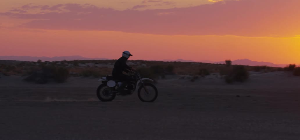 Person rides motorcycle across desert sands briefly popping a wheelie with sun shining from red sky on horizon
