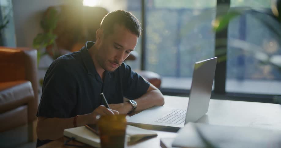 Man with mustache and blue shirt works on tablet and writes down in notebook at coffee table in living room at home