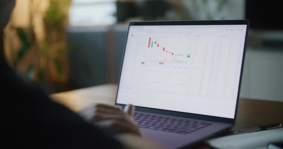 A man is sitting at a desk looking at a laptop computer with a graph on the screen