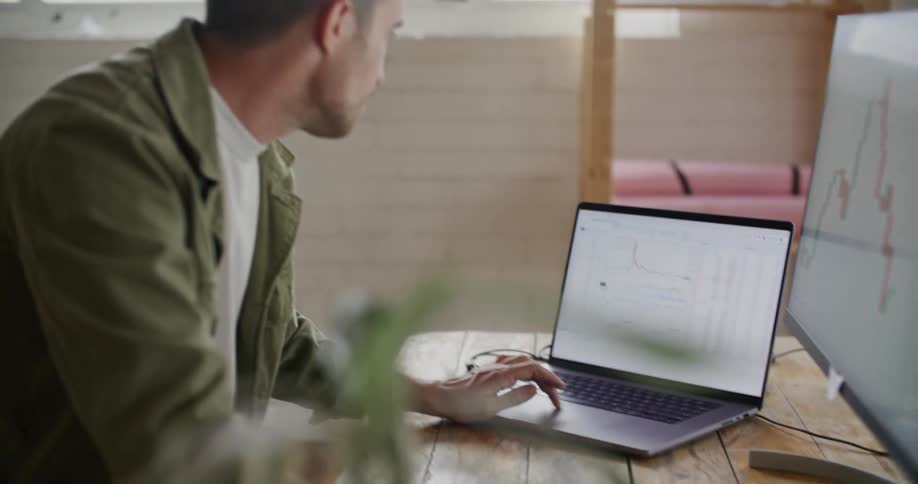 A man is sitting at a desk working on a laptop computer and looking at another monitor