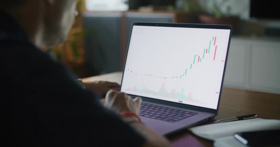 A man is sitting at a desk looking at a laptop computer with a graph on the screen