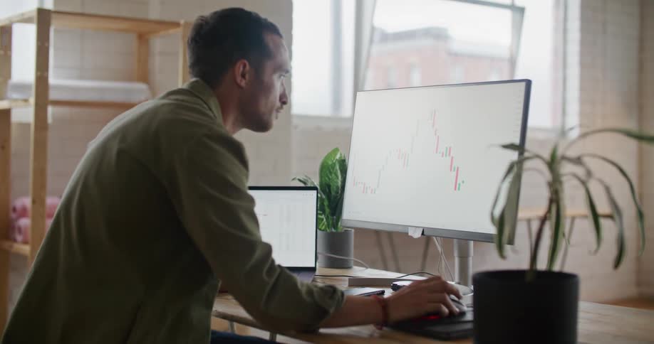 A man is sitting at a desk looking at a monitor with a graph on it and a laptop computer