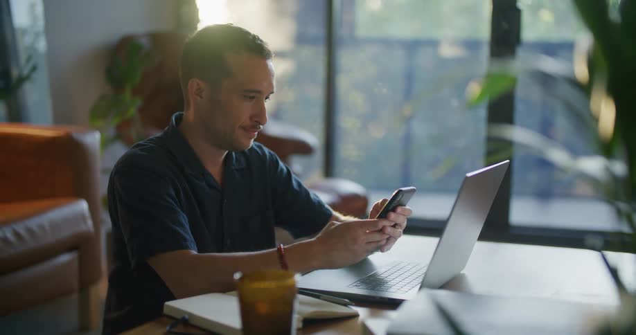 A man using a laptop at a low table stops to check his phone