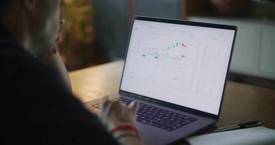 A man looks at stock charts on a laptop while sitting indoors