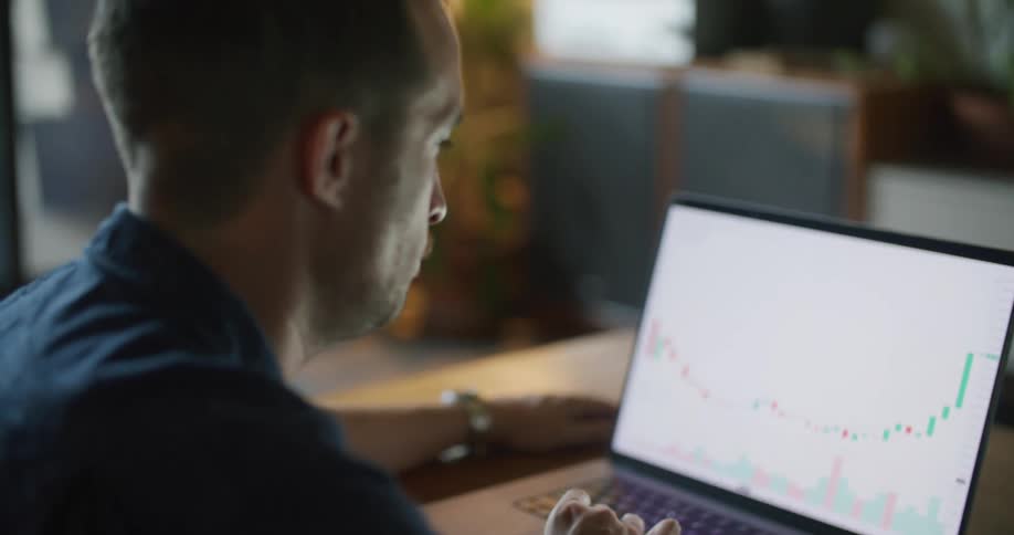 A man is sitting at a desk looking at a laptop computer with a graph on the screen