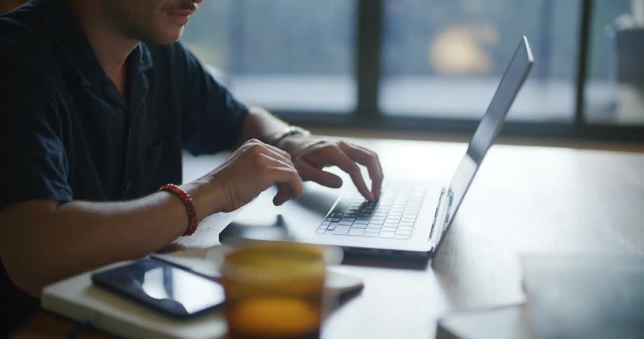 A man is sitting at a desk working on a laptop computer and writing down notes on paper