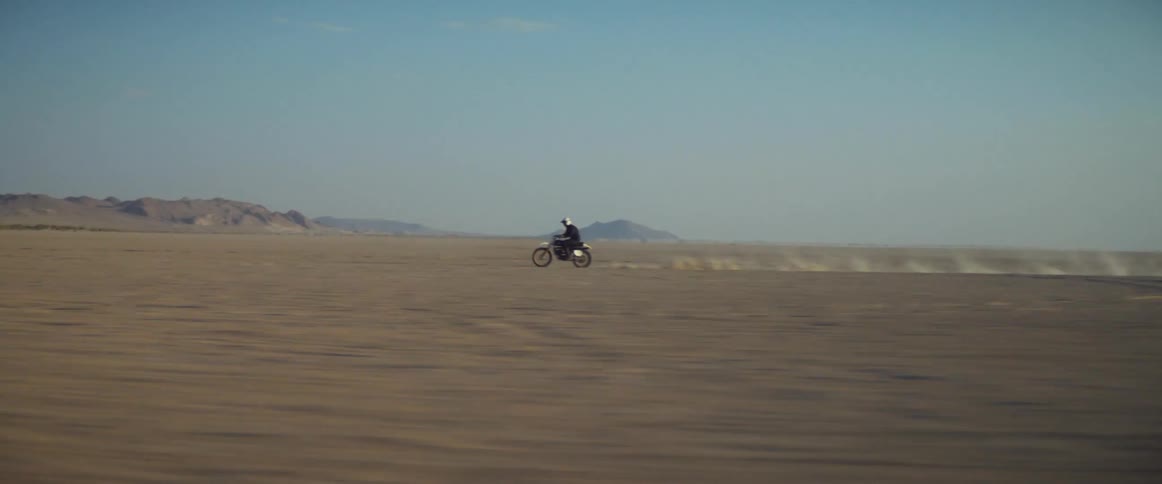 A cyclist wearing a helmet is riding a motorcycle across the desert with mountains in the distance