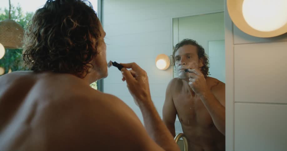 Shirtless man with wavy hair shaves himself looking in mirror in bathroom