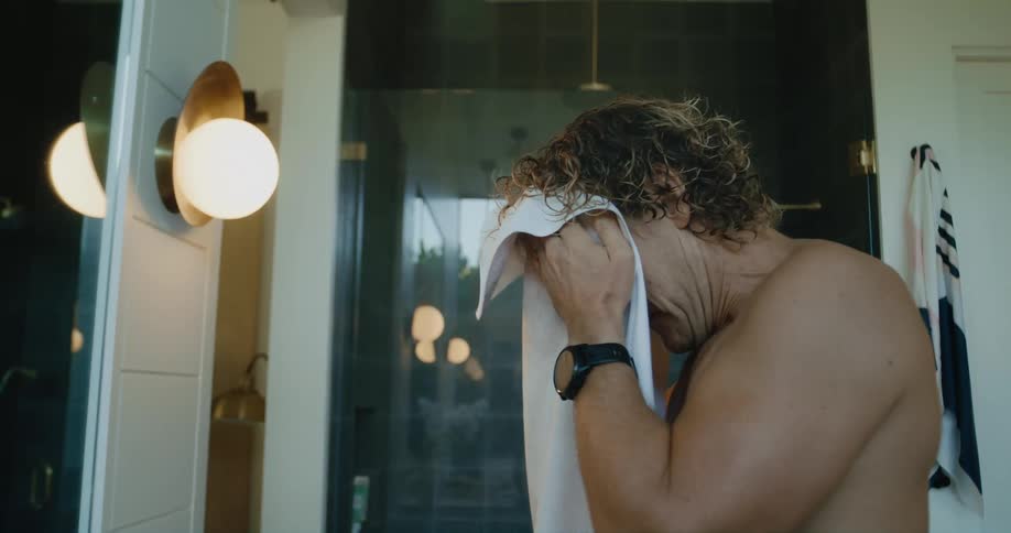 Shirtless man rubs a white towel on his face and neck while standing in a bathroom