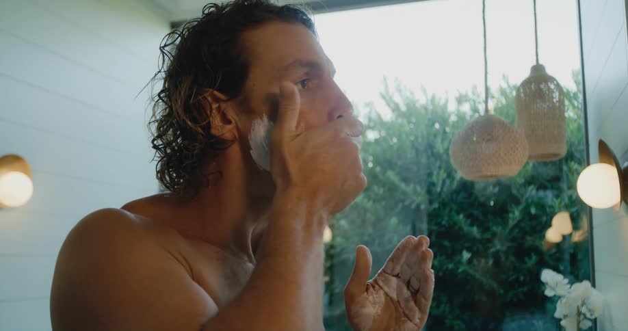 A shirtless man spreads shaving cream on his face while standing before a mirror in a bathroom during the day