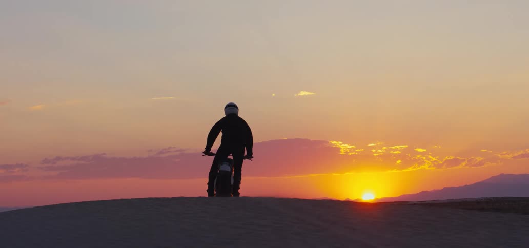 Person stands on motorcycle while riding it across sand toward sun on the horizon