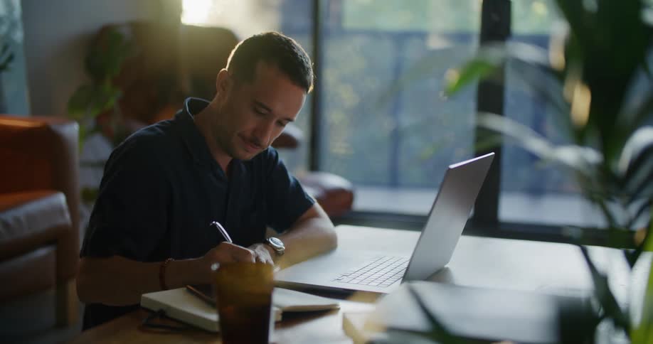 A man opens a laptop and begins to type before stopping to write in a notebook