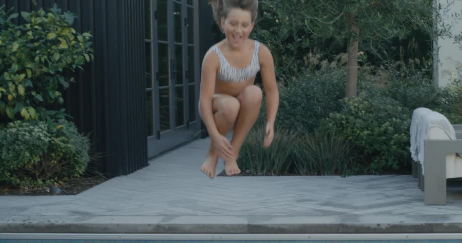 Girl wearing bikini runs along sidewalk and jumps into pool
