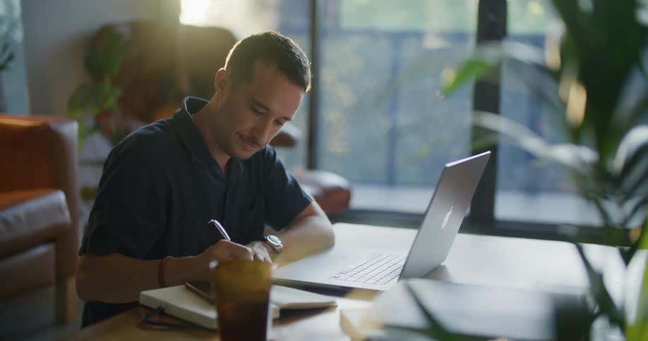 A man is sitting at a desk and opens a laptop computer and types on the keyboard then writes down notes on paper