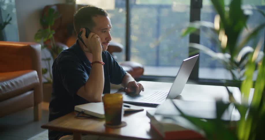 A man in an office makes a call on his cell phone as he uses his laptop